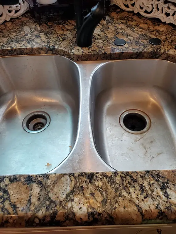 Stainless double kitchen sink with water spots, scratches and debris around both drains